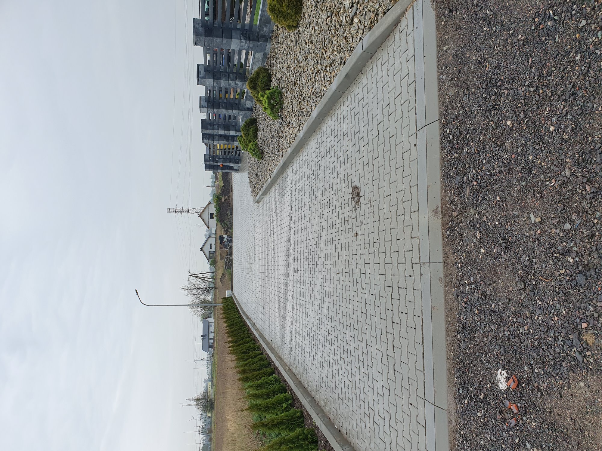 Szara ścieżka z kostki brukowej o wzorze puzzli, obrzeżona niskimi tujami i kamieniami ozdobnymi, prowadząca w stronę zabudowań mieszkalnych w pochmurny dzień.