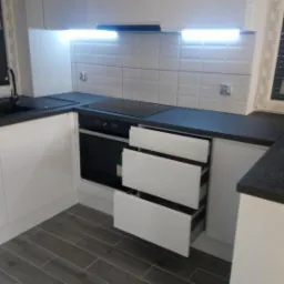 Nowoczesna, jasna kuchnia z białymi, błyszczącymi frontami szafek i ciemnym blatem, szare płytki podłogowe ułożone w jodełkę, dolne szuflady częściowo wysunięte.