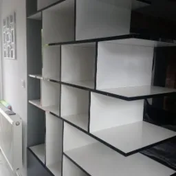 Nowoczesny regał na książki w kolorze białym i czarnym, z geometrycznymi półkami, ustawiony w jasnym wnętrzu, widoczny z boku.