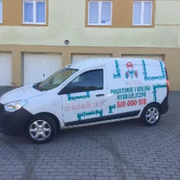 Biały samochód dostawczy z logo i numerem telefonu firmy hydraulicznej zaparkowany na brukowanym podjeździe przed budynkiem mieszkalnym z garażami.
