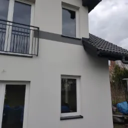 Wykończona elewacja domu jednorodzinnego z białym tynkiem, szarym pasem dekoracyjnym i ciemną stolarką okienną, widoczny fragment balkonu z metalową balustradą.