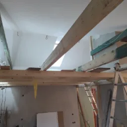 Poddasze w trakcie adaptacji: widoczne drewniane belki konstrukcyjne, częściowo otynkowane ściany, okno dachowe i drabina oparta o ścianę.