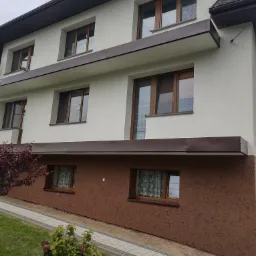 Dwukondygnacyjny dom z ocieploną elewacją w kolorze beżowym i brązowym, z ciemnobrązowymi ramami okiennymi i metalowymi elementami dekoracyjnymi, widok z poziomu ogrodu z młodym krzewem...