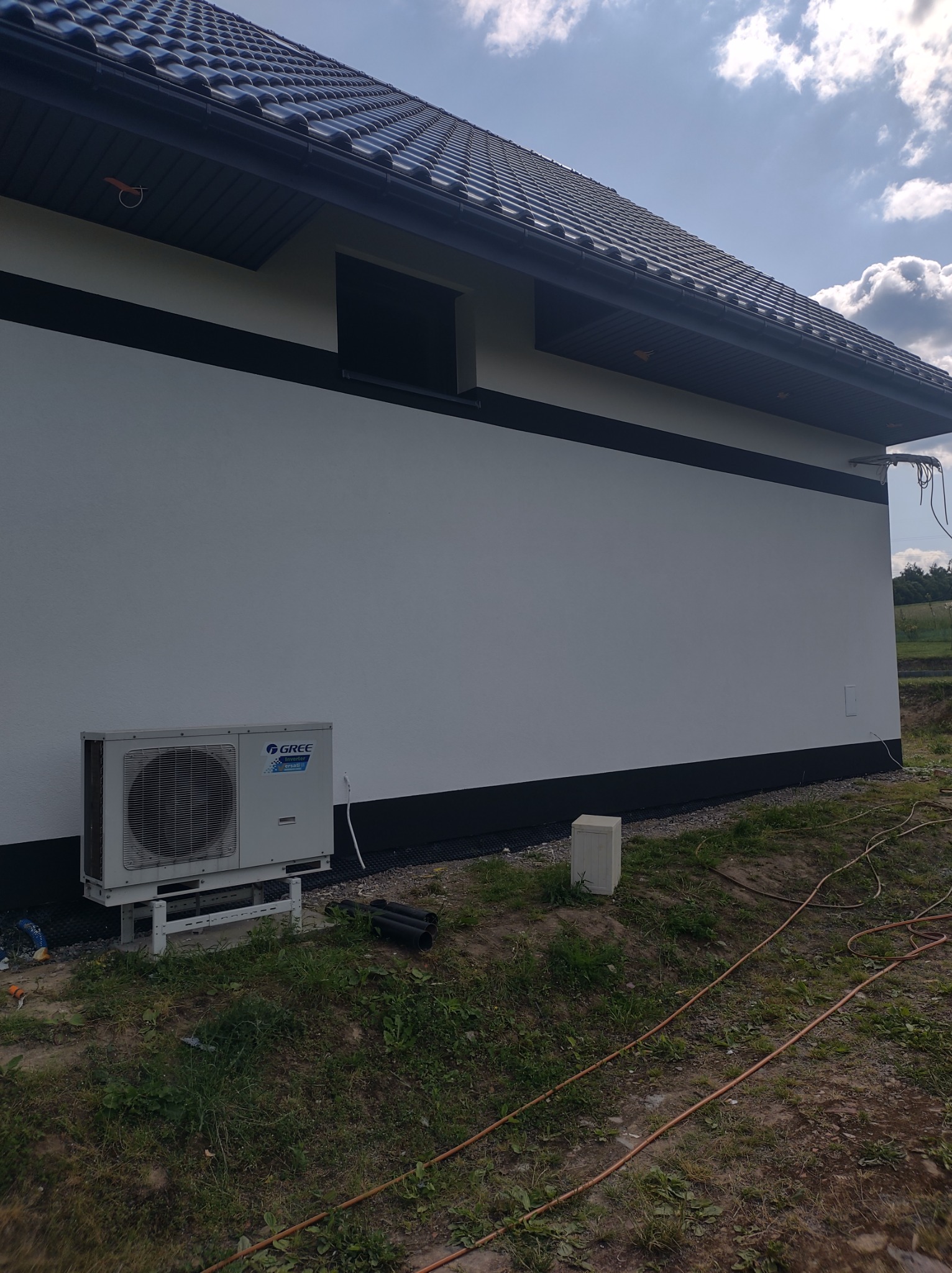 Biała ściana domu z czarnymi akcentami, widoczny klimatyzator Gree na zewnątrz, rury i kabel na ziemi, świeżo wykończona elewacja.