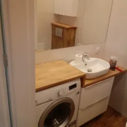 Umywalka nablatowa z baterią i akcesoriami, zamontowana na szafce z drewnianym blatem, obok pralka, w tle lustro i fragment drewnianej zabudowy toalety, całość w jasnej łazience wykończonej płytkami.