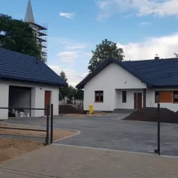 Nowoczesny dom jednorodzinny z garażem i czarnym ogrodzeniem panelowym, świeżo po ukończeniu prac elewacyjnych, z widoczną wieżą kościoła w tle.