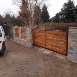 Drewniana brama wjazdowa z poziomymi deskami, osadzona na słupkach z szarego, dekoracyjnego betonu, obok zaparkowany biały samoch&oacute;d dostawczy.