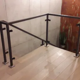 Szklana balustrada z ciemną, metalową ramą, mocowana do drewnianych schodów, z widocznymi uchwytami mocującymi szkło, na tle ściany z efektem rdzy.