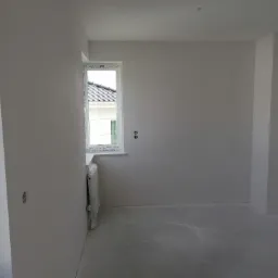 Wnętrze pokoju w trakcie prac przygotowawczych: świeżo nałożona biała gładź na ścianach i podłodze, okno z widokiem na dach domu, zakryty grzejnik.