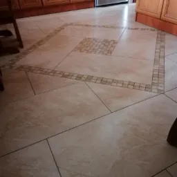 Beżowa, kwadratowa podłoga z płytek ceramicznych w kuchni, z dekoracyjnym wzorem mozaiki w kształcie kwadratu i obramowania z małych płytek, widoczne brązowe szafki kuchenne i fragment krzesła.