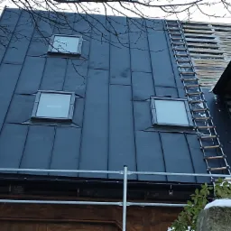 Nowe, ciemne poszycie dachu z trzema oknami połaciowymi, częściowo zdemontowane stare pokrycie widoczne z boku, drabina dekarska oparta o dach.