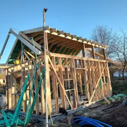 Wznoszenie szkieletu drewnianego domu na fundamencie, widoczne elementy konstrukcyjne dachu i ściany, w tle drzewa i błękitne niebo.