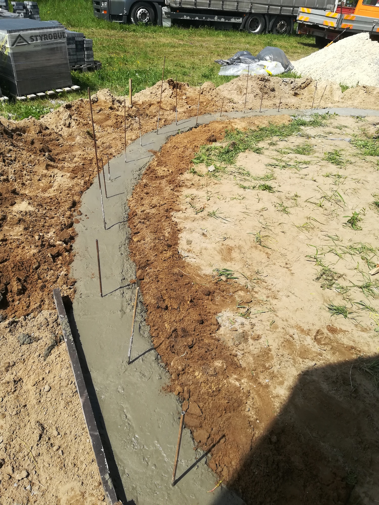 Wykopany rów z wylewką betonową, wzmocniony prętami, widoczne palety kostki brukowej i ciężarówka w tle.