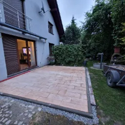 Wyłożony kostką taras przy domu jednorodzinnym, obrzeża z szarego betonu, widoczny fragment przyczepki samochodowej i bujna roślinność w tle.