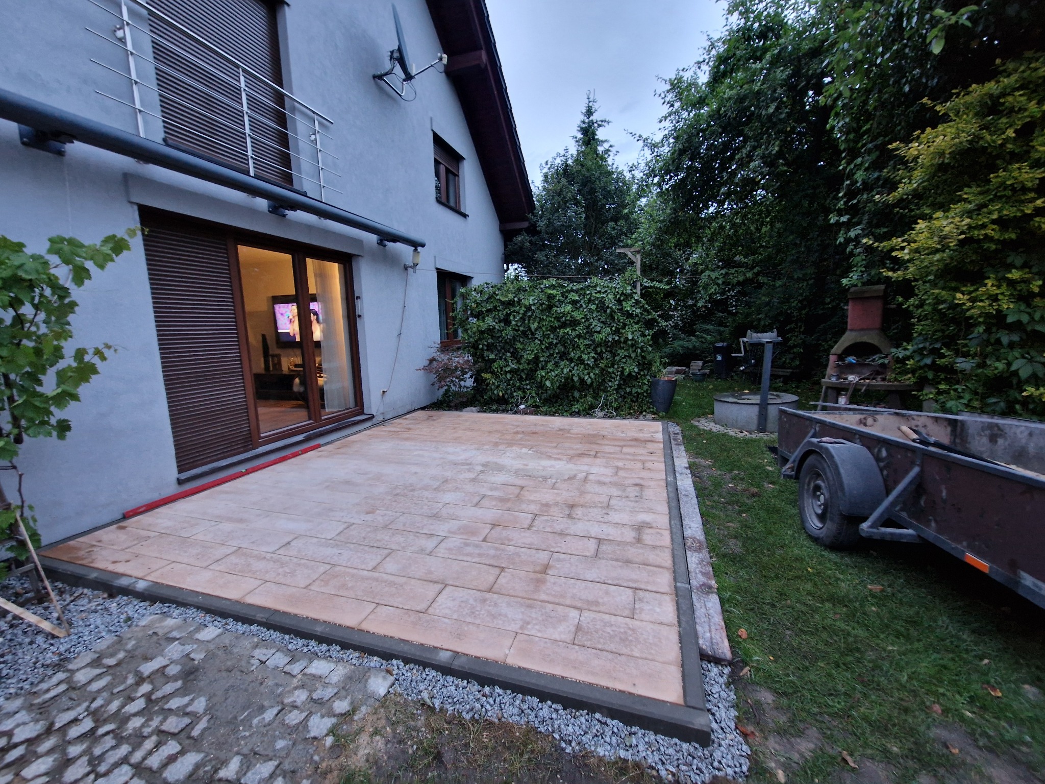 Wyłożony kostką taras przy domu jednorodzinnym, obrzeża z szarego betonu, widoczny fragment przyczepki samochodowej i bujna roślinność w tle.
