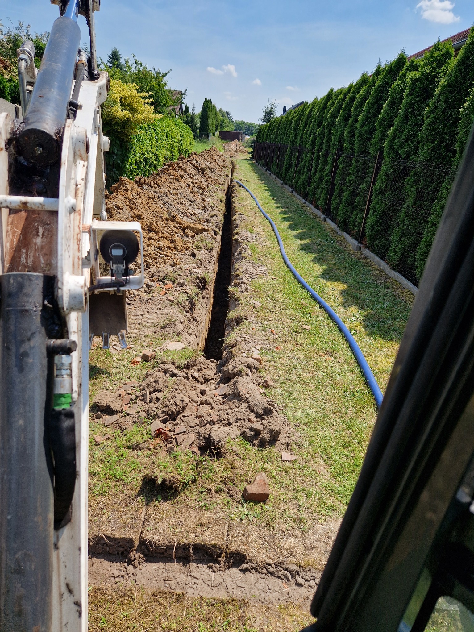 Wąski wykop w ziemi, widok z kabiny koparki, obok zielony trawnik i rząd tuj, niebieski wąż leży wzdłuż wykopu.