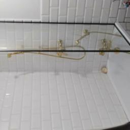 Dawid Rządkowski - Nowoczesna kabina prysznicowa z czarną ramą i złotą armaturą na tle białych, podłużnych płytek. W rogu widoczna dekoracyjna ściana z mozaiką.