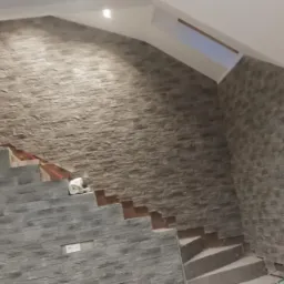 Ściana z dekoracyjną okładziną imitującą kamień, fragment schodów w trakcie wykończenia, widoczne niedokończone stopnie i elementy montażowe.