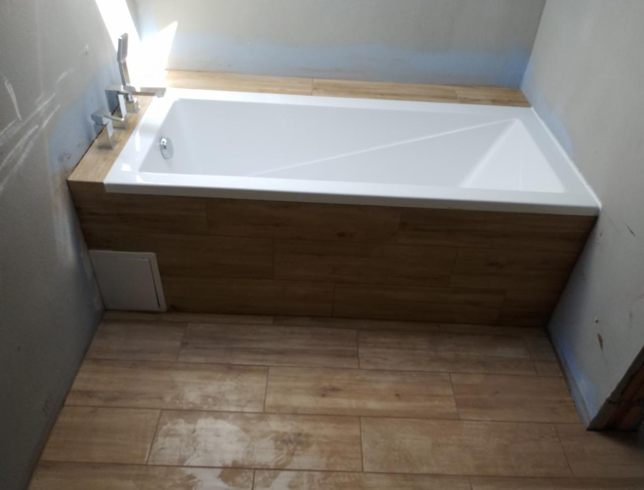 Biała wanna obudowana panelami imitującymi drewno, z dopasowaną kolorystycznie podłogą z płytek, widoczna armatura i fragment ściany w trakcie prac wykończeniowych.