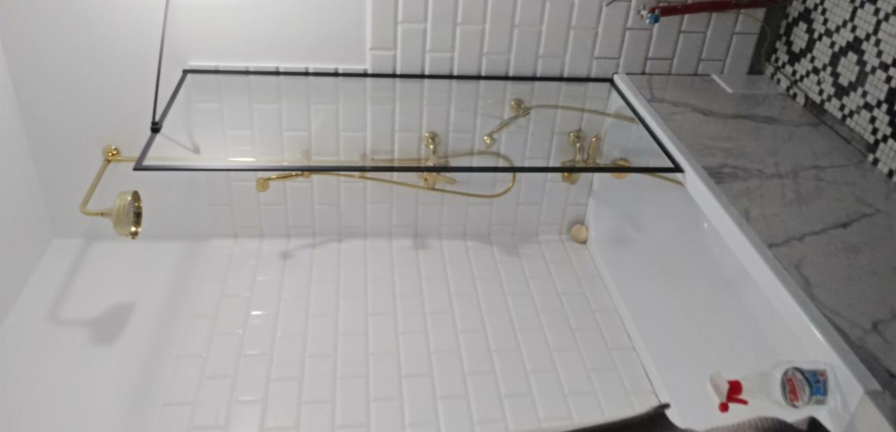 Nowoczesna kabina prysznicowa z czarną ramą i złotą armaturą na tle białych, podłużnych płytek. W rogu widoczna dekoracyjna ściana z mozaiką.