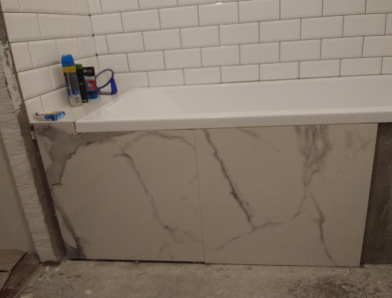 Obudowa wanny wykończona płytkami imitującymi marmur, białe płytki metra na ścianie, widoczne materiały budowlane i narzędzia, etap prac wykończeniowych.