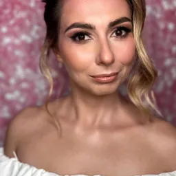 Portret kobiety z delikatnym makijażem i upiętymi włosami na tle różowej ściany. Ubrana w białą sukienkę z odkrytymi ramionami, z lekkim uśmiechem.