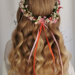 HD Make Up Hanna Durczak - Blond włosy z lokami, ozdobione wiankiem z polnych kwiatów i wstążkami w odcieniach różu i pomarańczy na tle białej tkaniny.
