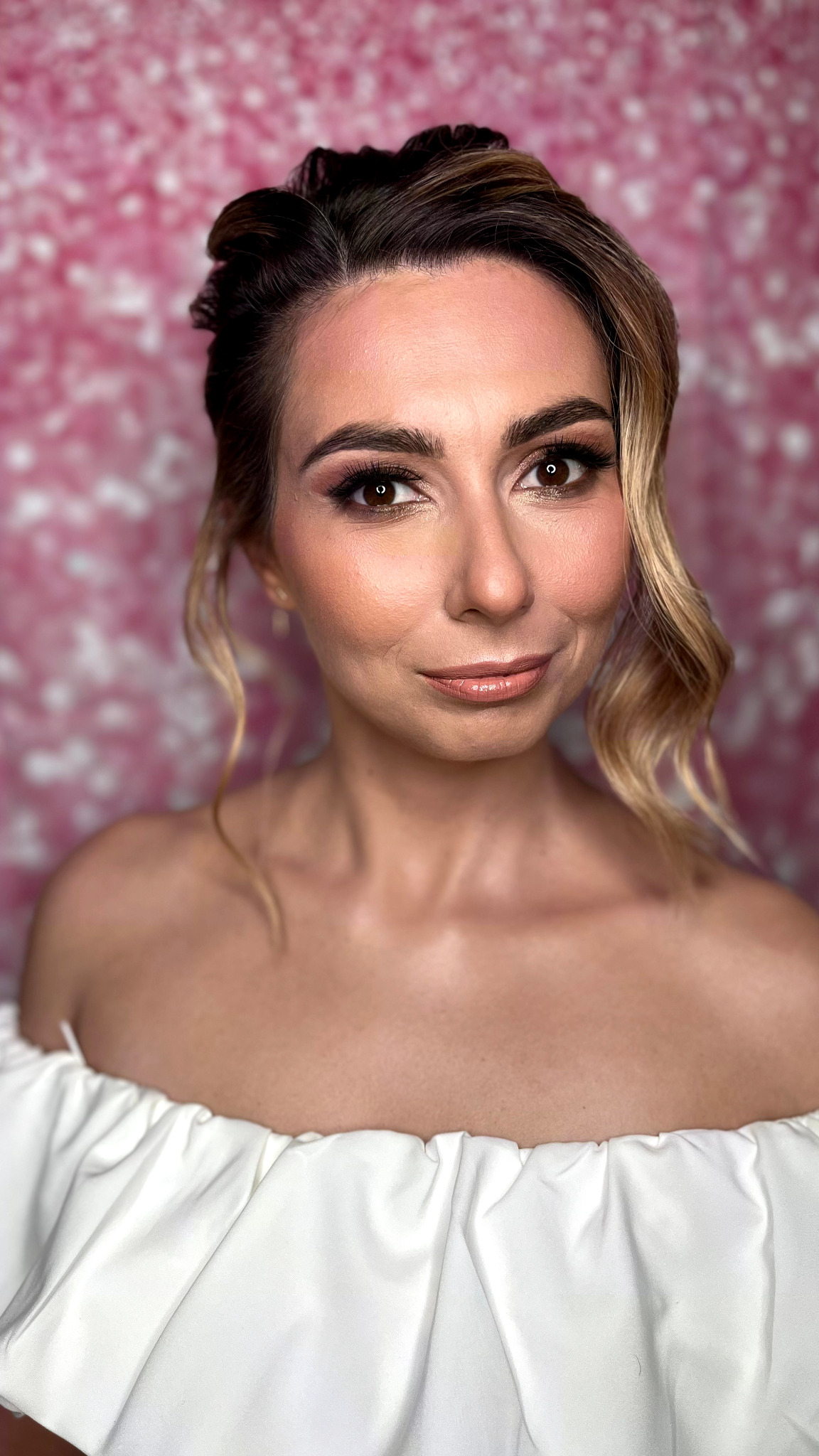 Portret kobiety z delikatnym makijażem i upiętymi włosami na tle różowej ściany. Ubrana w białą sukienkę z odkrytymi ramionami, z lekkim uśmiechem.