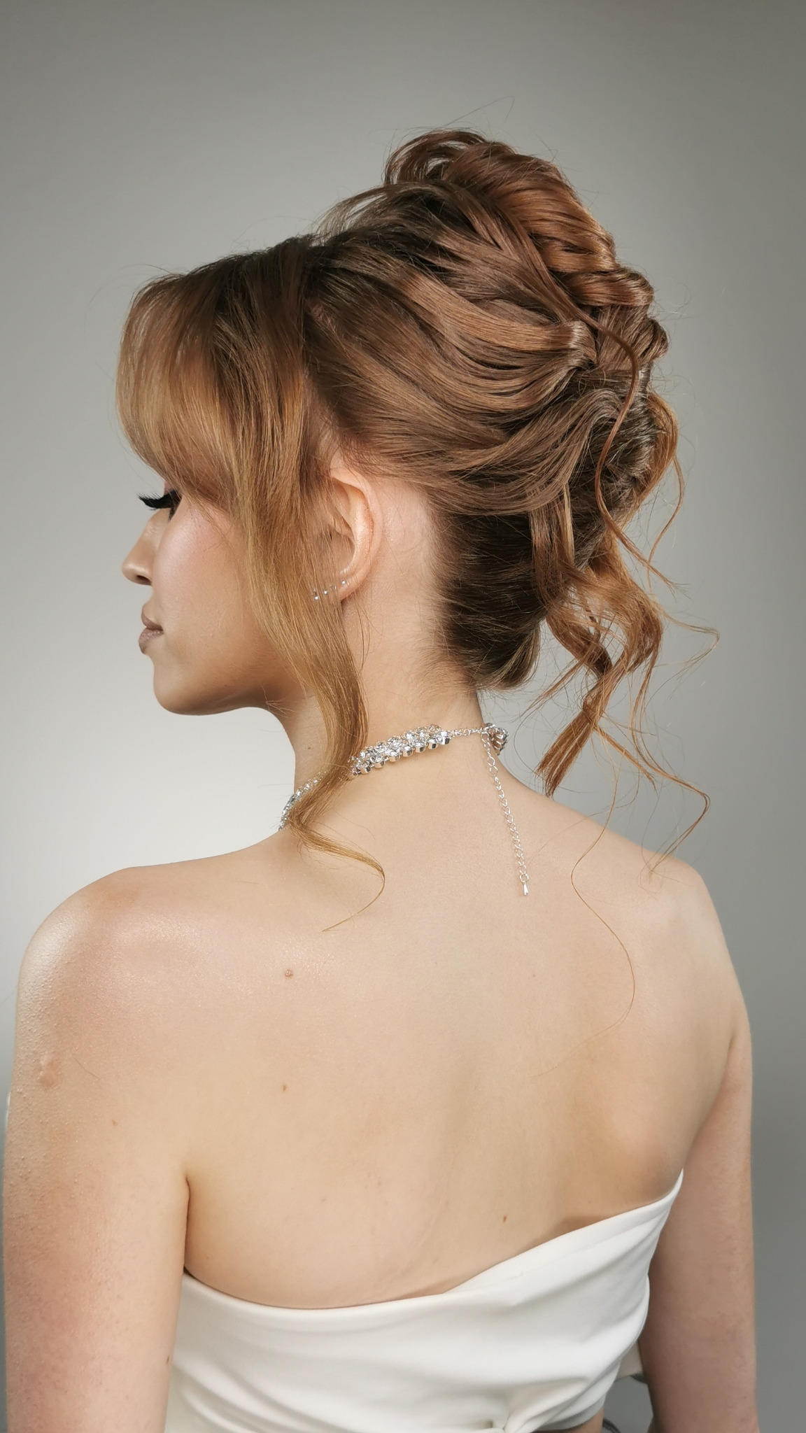 Elegancka fryzura upięta z rudych włosów, widok z tyłu na szyję z biżuterią, subtelny profil kobiety. Stylizacja ślubna.