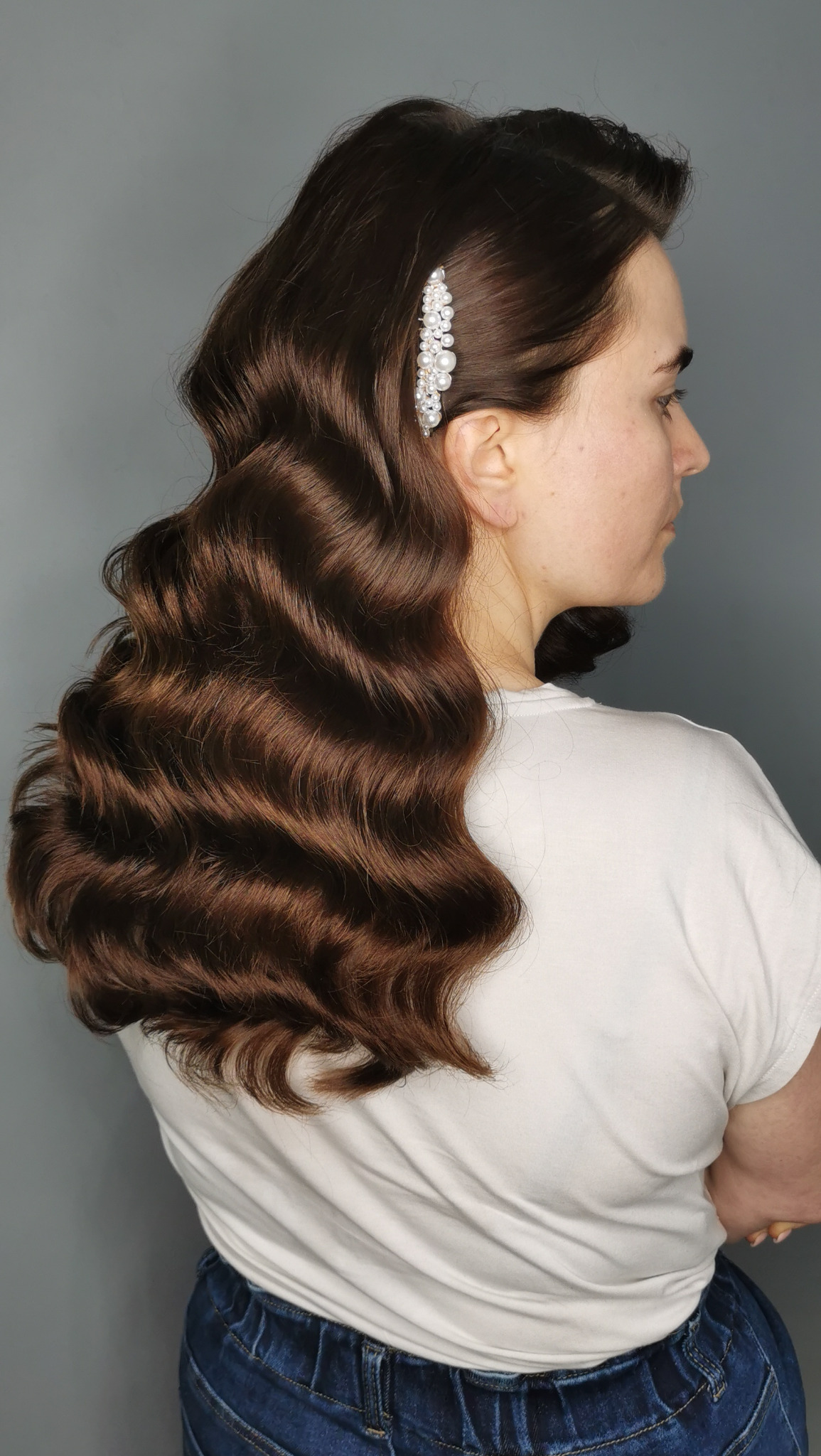 Brunetka z upięciem w stylu retro, fale i ozdobna spinka z perełkami. Profil twarzy, neutralne tło. Elegancka fryzura na specjalną okazję.