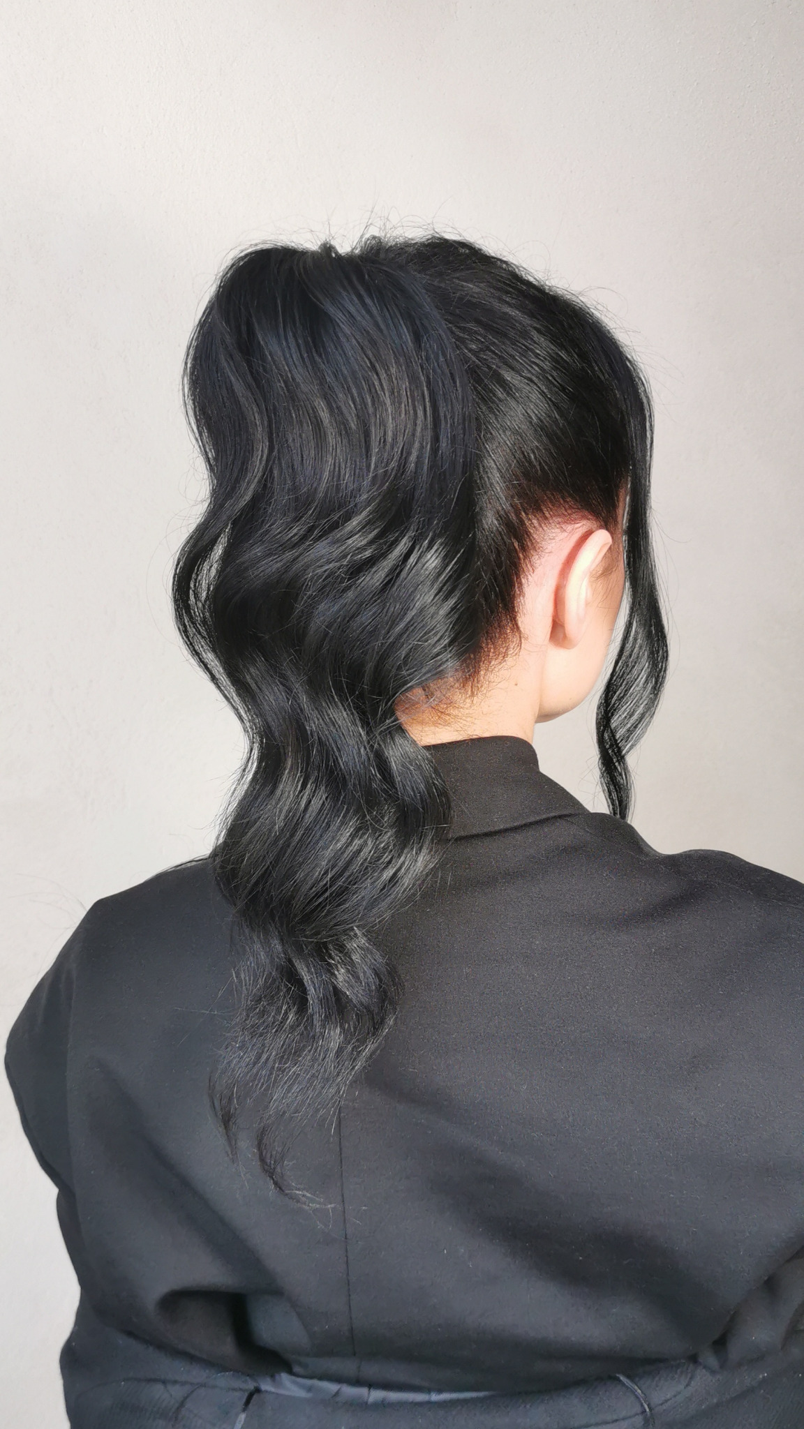Elegancka fryzura upięta w wysoki kucyk z pofalowanymi pasmami, widok z tyłu, czarne włosy, gładko zaczesane u nasady, ciemna marynarka, jasne tło.