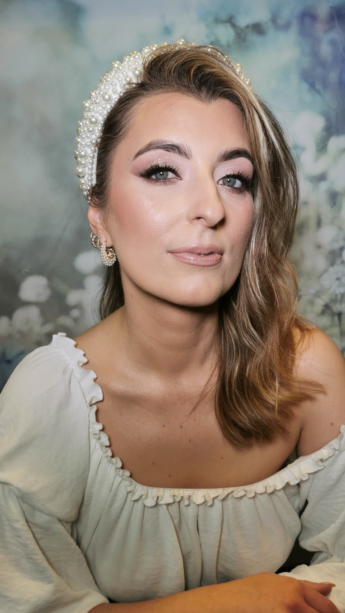 Portret kobiety z profesjonalnym makijażem, w perłowej opasce i kolczykach, na tle abstrakcyjnym. Delikatny makijaż podkreśla urodę.