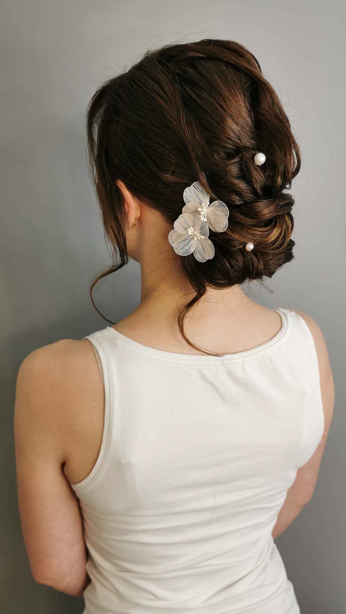 Upięcie ślubne z kwiatowymi ozdobami i perłami, widok z tyłu na brunetkę w białej koszulce na tle szarej ściany. Subtelna elegancja.
