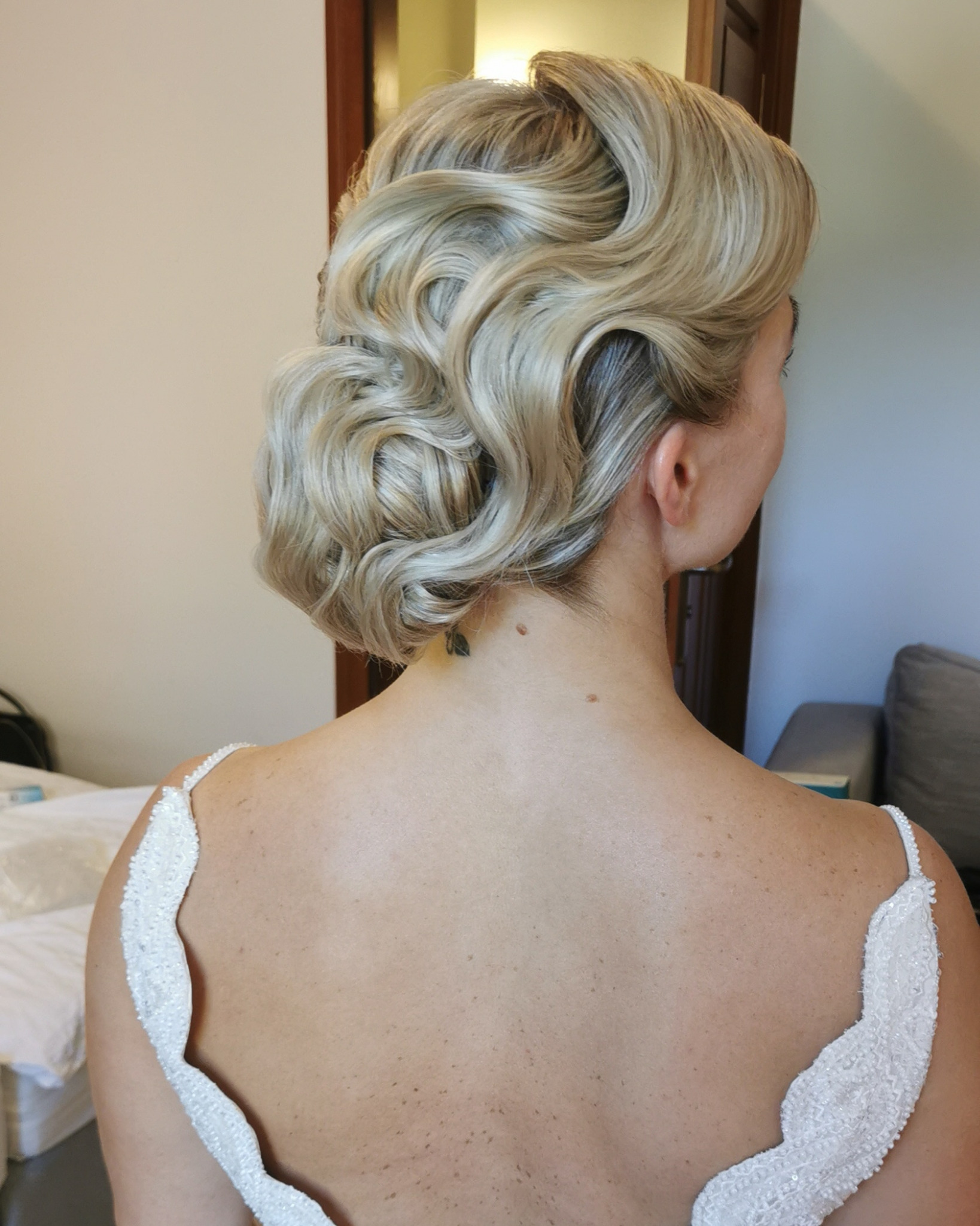 Elegancka upięta fryzura blond z tyłu głowy kobiety w sukni ślubnej, widoczny tatuaż na karku i fragment pokoju.