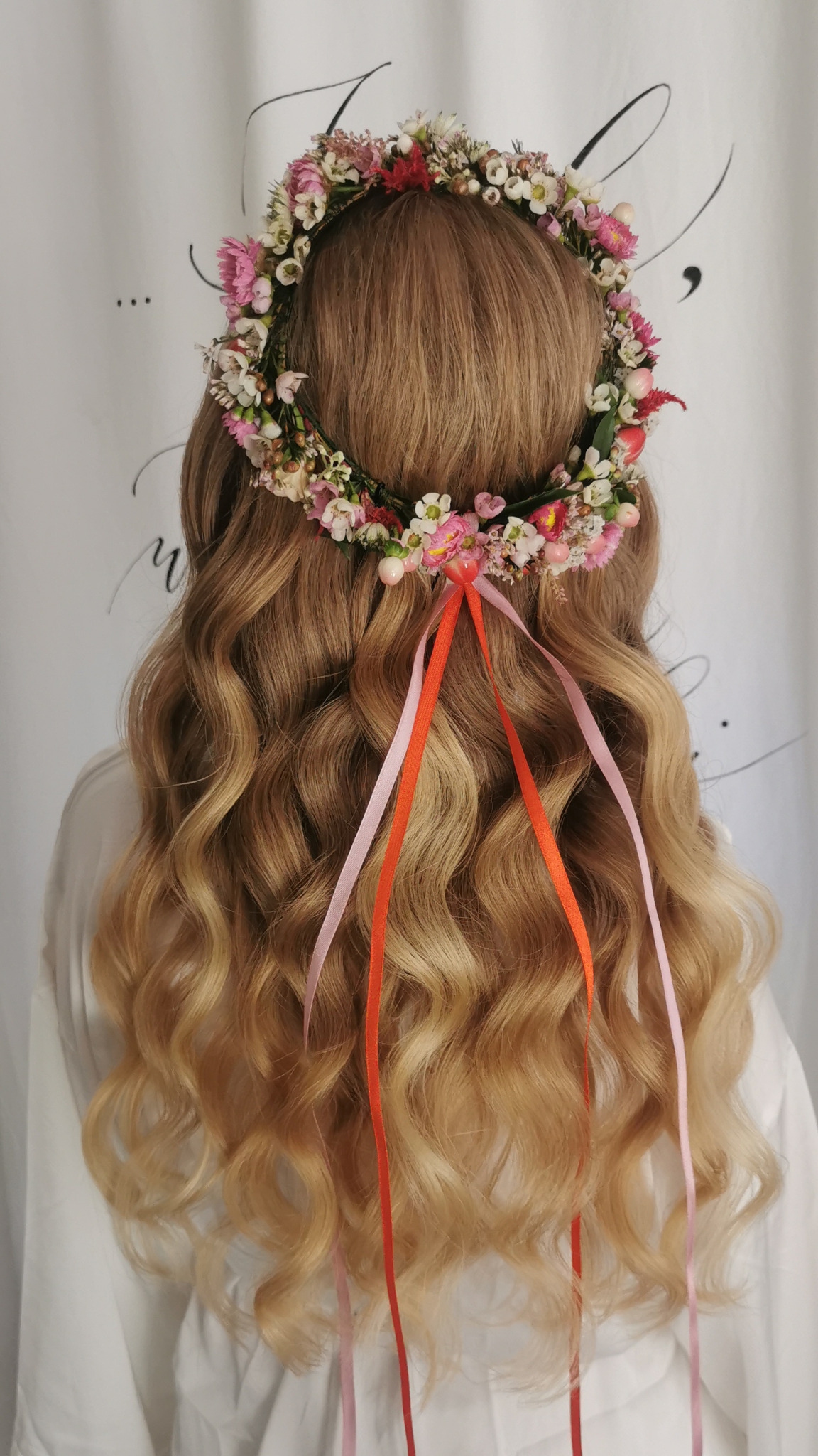 Blond włosy z lokami, ozdobione wiankiem z polnych kwiatów i wstążkami w odcieniach różu i pomarańczy na tle białej tkaniny.