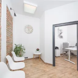 Jasne wnętrze gabinetu lekarskiego z ceglaną ścianą, białymi krzesłami i widokiem na biurko w drugim pomieszczeniu.