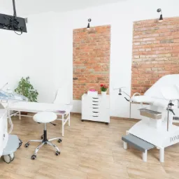 Jasne wnętrze gabinetu lekarskiego z kozetką, fotelem ginekologicznym marki Tomedic, aparaturą USG i ścianami z elementami dekoracyjnej cegły.