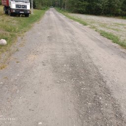 Wodrol Krzysztof Książek - Długa, szutrowa droga gruntowa z widocznymi koleinami, prowadząca przez teren zielony w kierunku lasu, z zaparkowaną ciężarówką MAN po lewej stronie i widocznym kamieniem obok ogrodzenia.