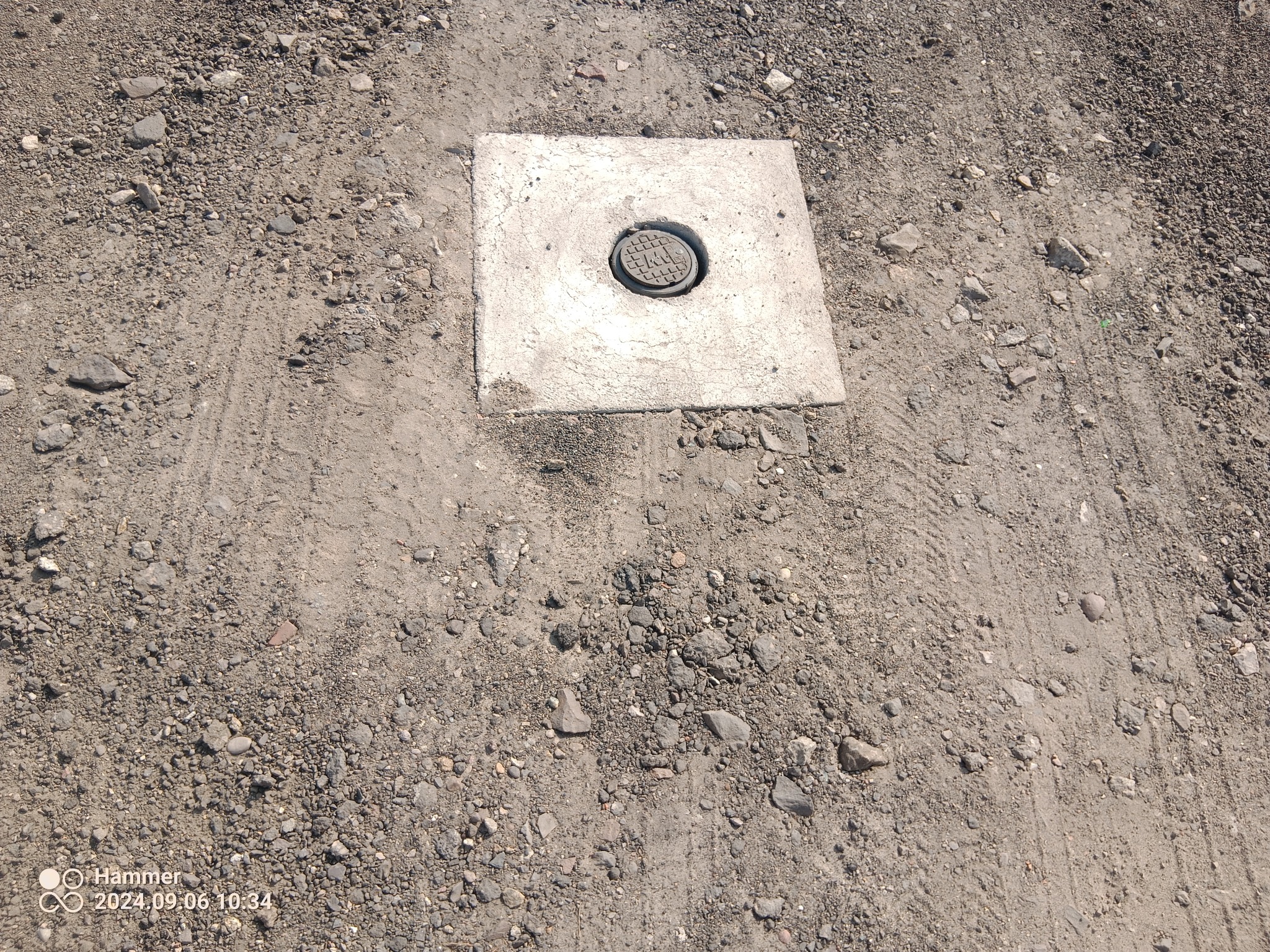 Betonowa studzienka rewizyjna z metalową pokrywą, wkomponowana w grunt z drobnym żwirem i piaskiem, widoczna z góry.