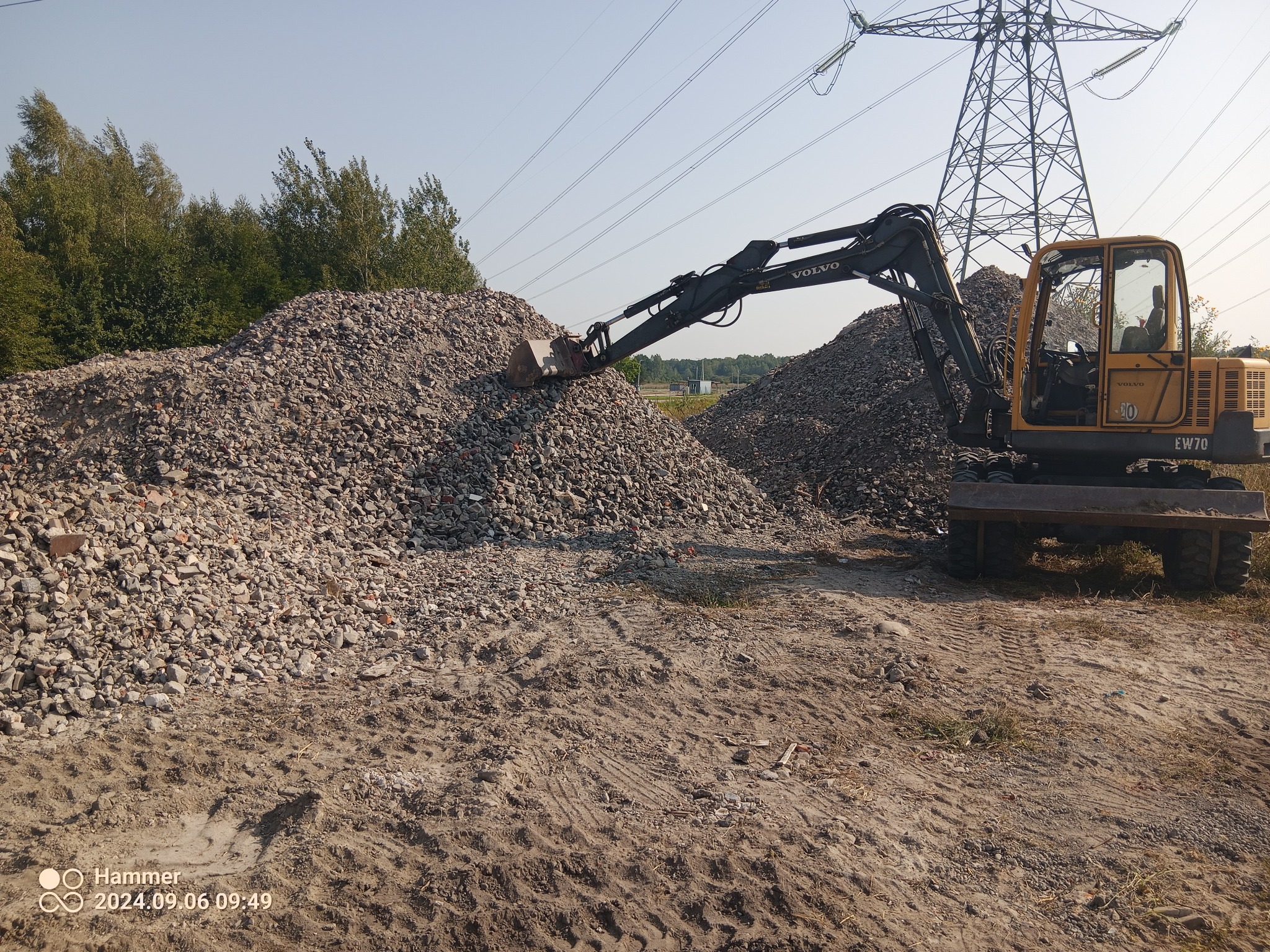 Żółta koparka Volvo EW70 nabiera łyżką gruz z hałdy na placu budowy, w tle las i słupy wysokiego napięcia.