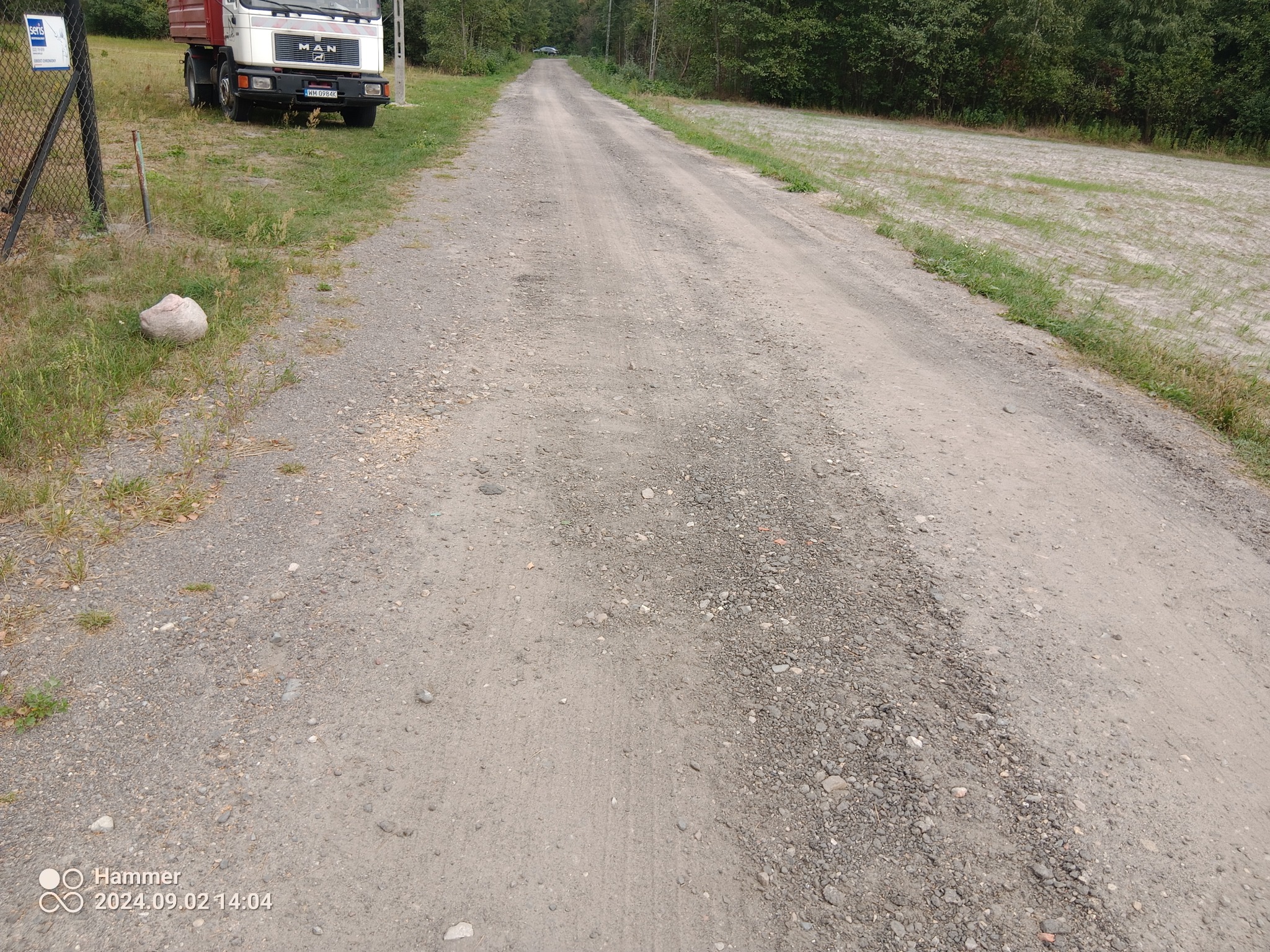 Długa, szutrowa droga gruntowa z widocznymi koleinami, prowadząca przez teren zielony w kierunku lasu, z zaparkowaną ciężarówką MAN po lewej stronie i widocznym kamieniem obok ogrodzenia.