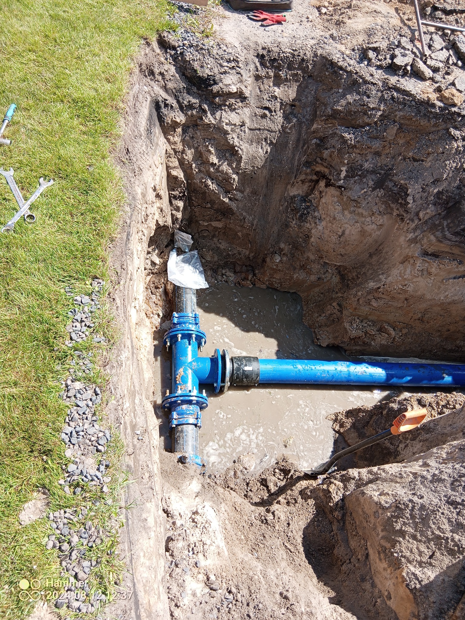 Wykonany wykop z częściowo zamontowanym, niebieskim przyłączem wodociągowym, wypełniony wodą, widoczna ziemia, trawa i narzędzia.