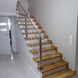 Drewniane schody z metalową balustradą na białych wspornikach, pionowe pasy na ścianie, widok z dołu.