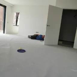 Mężczyzna w niebieskiej koszulce leży na podłodze zabezpieczonej folią, w trakcie prac wykończeniowych w jasnym, przestronnym wnętrzu z białymi ścianami i otwartymi drzwiami, widoczny poziomica...