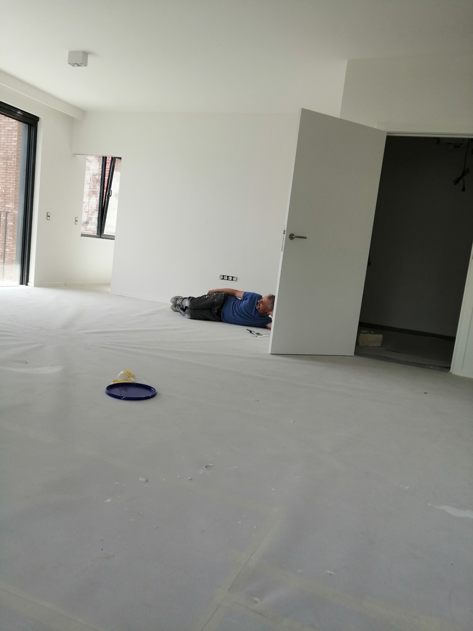 Mężczyzna w niebieskiej koszulce leży na podłodze zabezpieczonej folią, w trakcie prac wykończeniowych w jasnym, przestronnym wnętrzu z białymi ścianami i otwartymi drzwiami, widoczny poziomica...