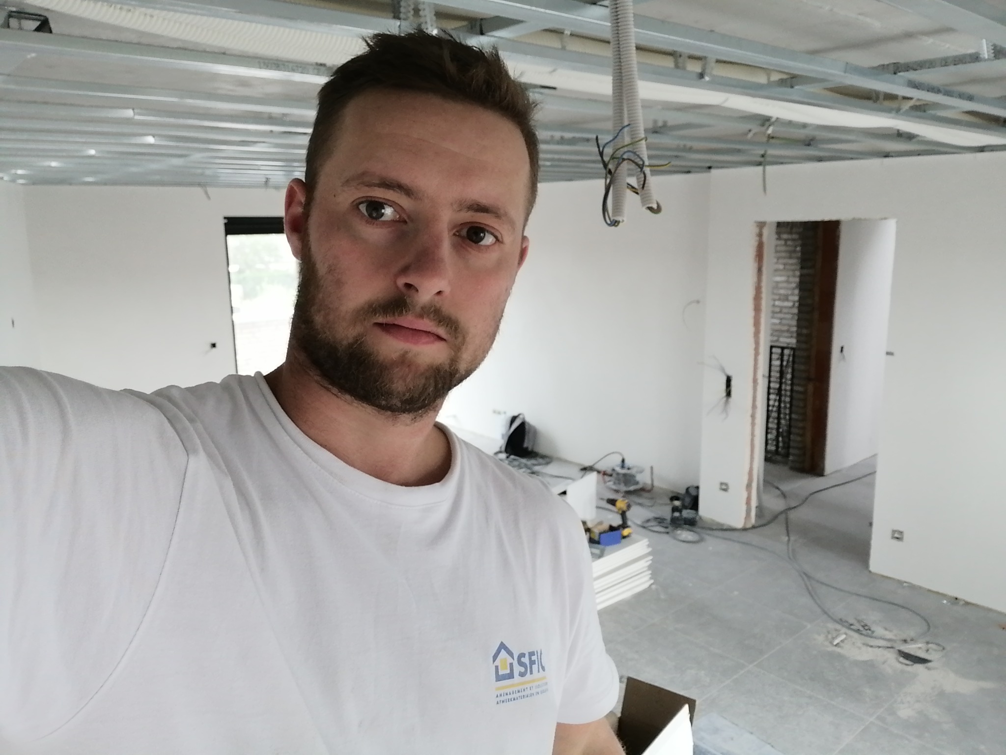 Mężczyzna w białej koszulce z logo firmy na tle surowego wnętrza w trakcie remontu: widoczne profile sufitowe, kable, narzędzia i płyty gipsowo-kartonowe.