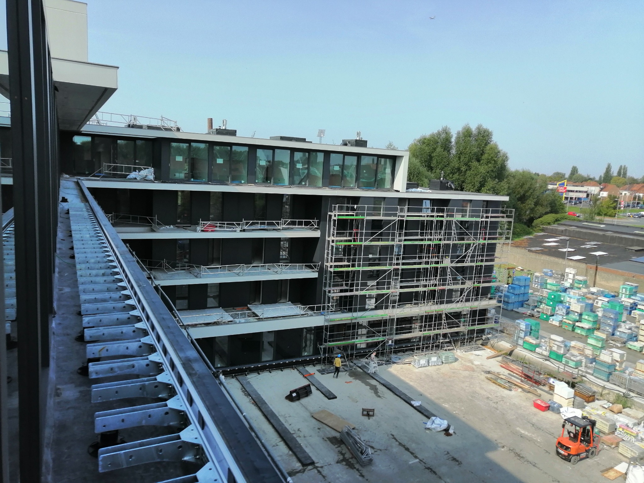 Wysoki budynek w trakcie remontu z rusztowaniami, widoczne materiały budowlane, robotnicy w oddali i pomarańczowy wózek widłowy na placu budowy.