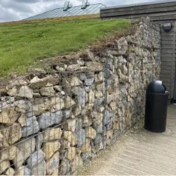 Gabionowa ściana oporowa z kamienia naturalnego, porośnięta trawą na dachu budynku, obok wejścia z czarnym koszem na śmieci.