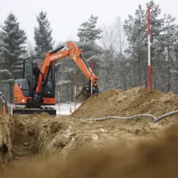 Pomarańczowa koparka Hitachi 33U w trakcie wykopu wąskiego rowu w ziemi, widoczne ułożone szare rury, w tle las w zimowej scenerii.
