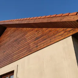 Nowe drewniane podbitki dachowe z brązową bejcą i pomarańczową dachówką ceramiczną, widoczne z dołu na tle błękitnego nieba.